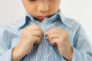 cute asian boy getting dressed for school, buttoning long sleeved shirt with concentration and happiness to go back to school