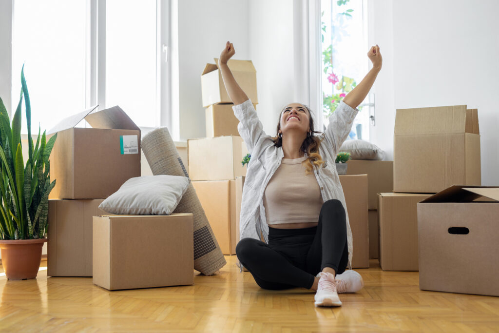 young woman sitting on floor in new apartment with boxes and raising arms in joy