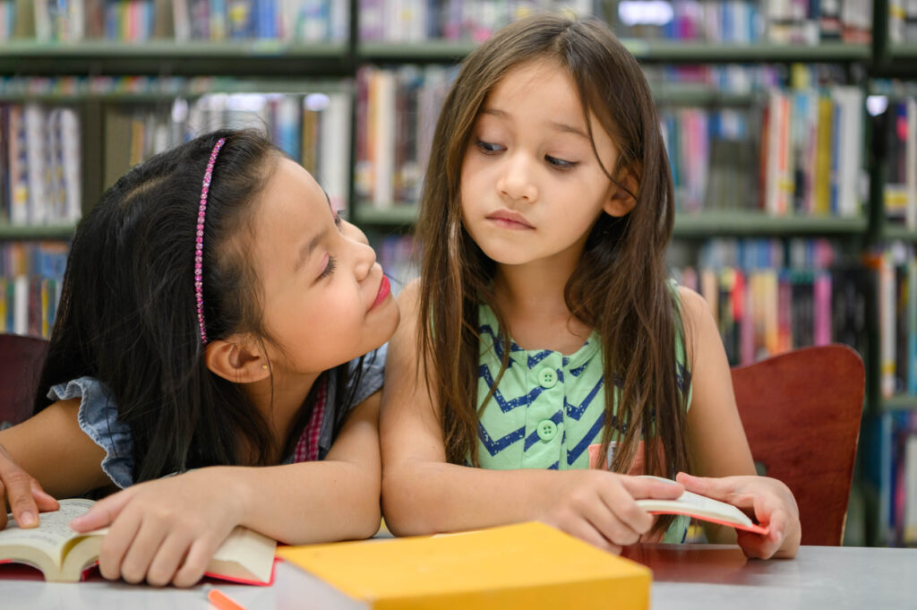 two cute girls are jealous of each other while reading books in library while teacher teaching. people lifestyles and education. young friendship and kids relationship in school concept. daycare theme