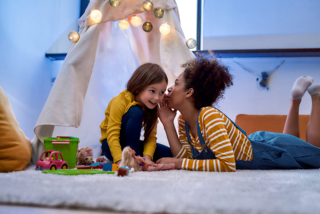 african american woman baby sitter entertaining caucasian cute little girl. they are gossiping and telling secrets sitting in kids room