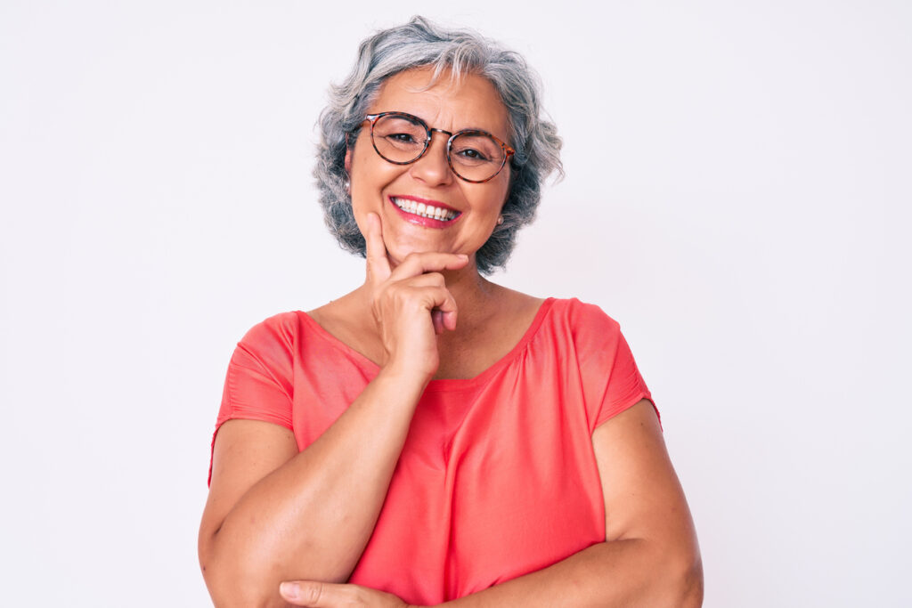 senior hispanic grey haired woman wearing casual clothes and glasses looking confident at the camera with smile with crossed arms and hand raised on chin. thinking positive.