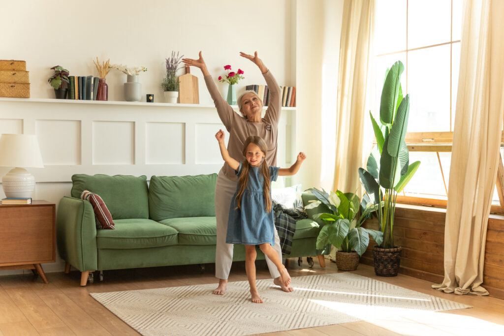 energetic senior female grandma dancing together with granddaughter in living room.