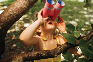 closeup portrait of little girl looking through a binoculars searching for an imagination or exploration in summer day in park. child playing with binocular pretend safari game outdoors in the forest