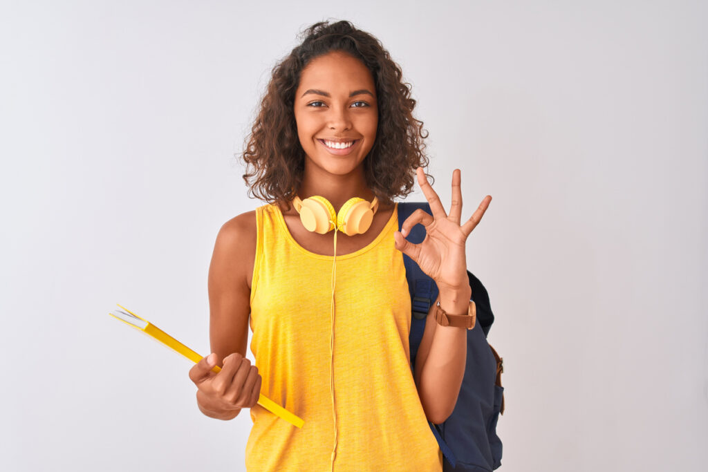 brazilian student woman wearing backpack holding notebook over isolated white background doing ok sign with fingers, excellent symbol