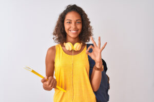 brazilian student woman wearing backpack holding notebook over isolated white background doing ok sign with fingers, excellent symbol
