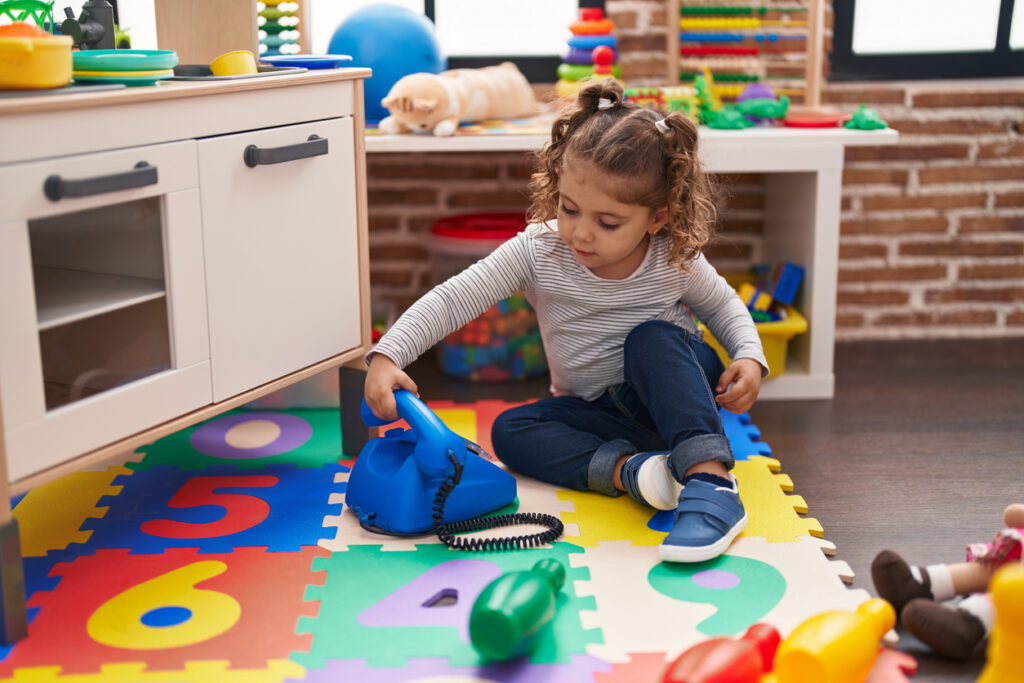 adorable caucasian girl playing with telephone toy sitting on floor at kindergarten