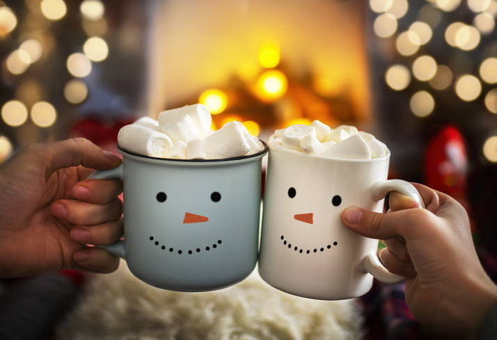 mom and child are relaxing together on a cozy winter evening by the fireplace, close up of two hands with snowman face cup of hot cocoa with marshmallows. christmas holidays, happy moments at home.