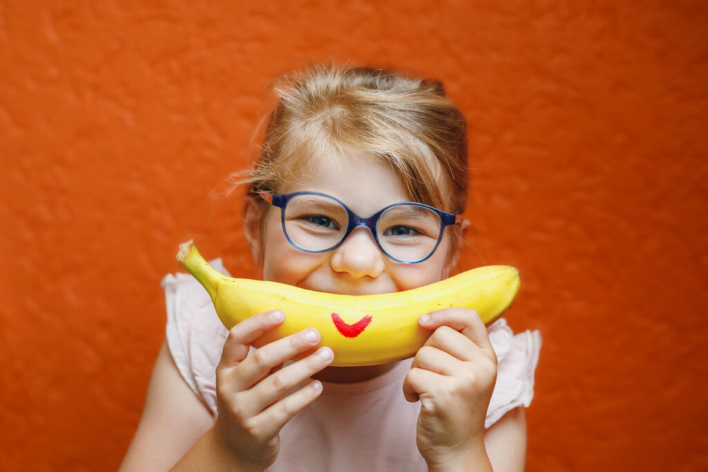 happy little child girl with yellow banana like smile on orange background. preschool girl with glasses smiling. healthy fruits for children