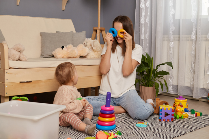 activities with children at home, early childhood development. young adult woman sitting on floor in living room, playing with her baby child with pyramid, covering her eyes with rings, having fun.